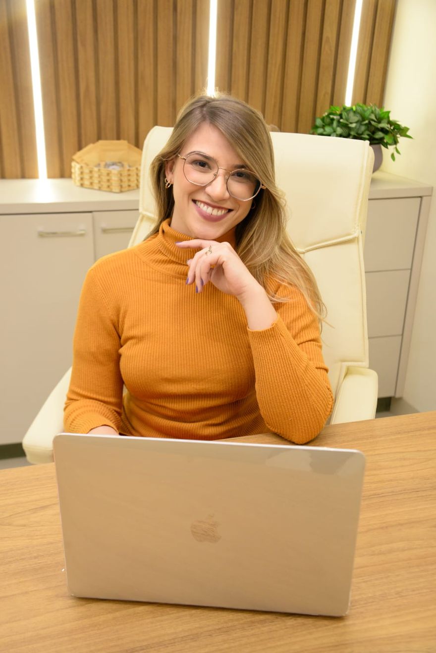 Dra. Gabriela Figueiredo sorrindo em seu consultório, sentada diante do computador